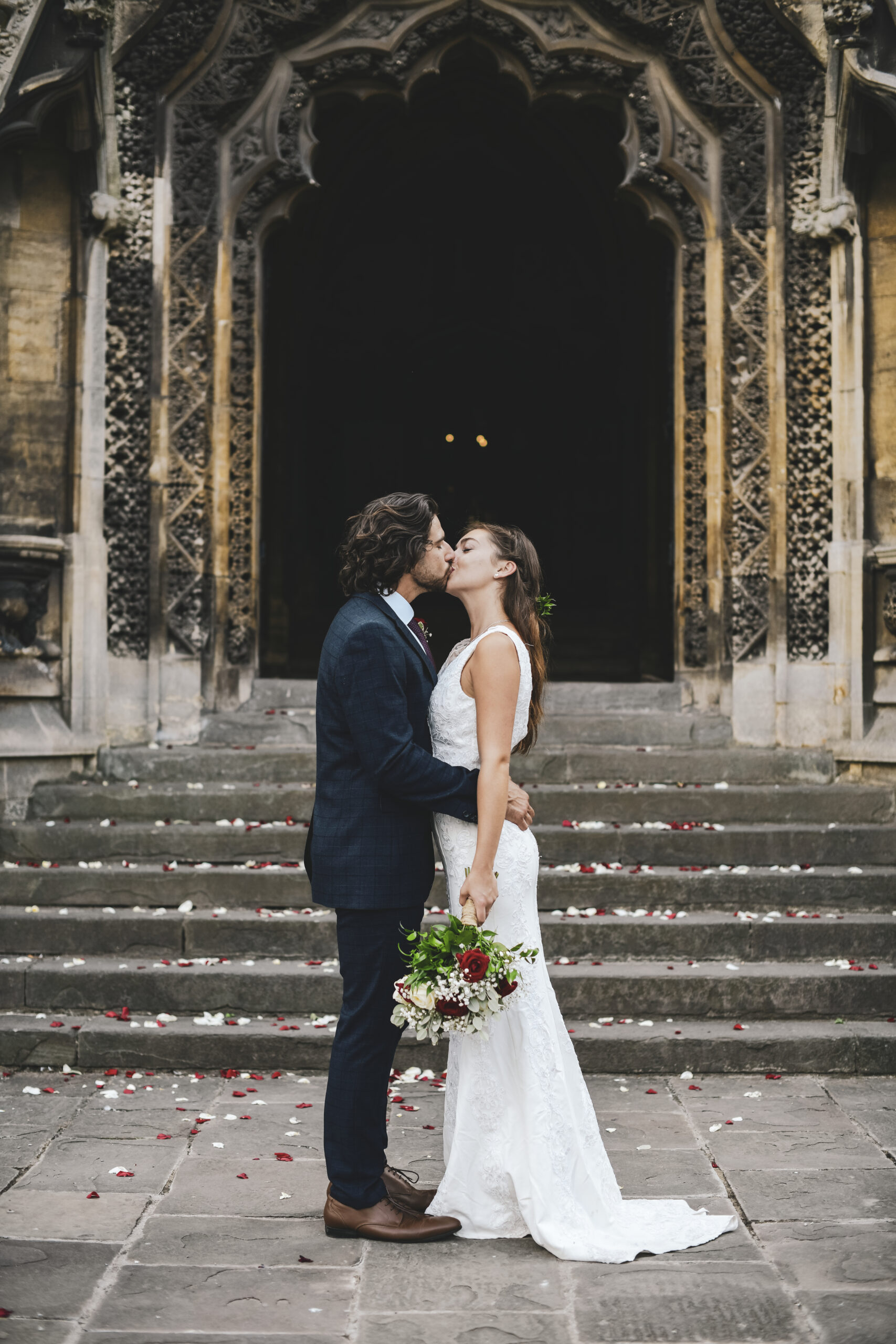 Brautpaar küsst sich vor einer Kirche, die braut hat einen Rot weis grünen Brautstraus und auf dem Boden liegt Konfetti verteilt, Jessis Weddings